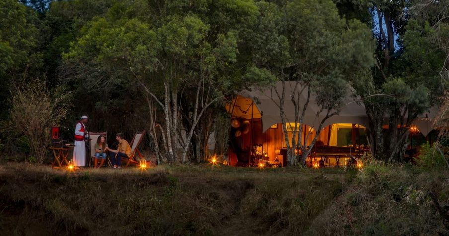 A tented camp lit by lanterns at dusk, with guests seated by the trees