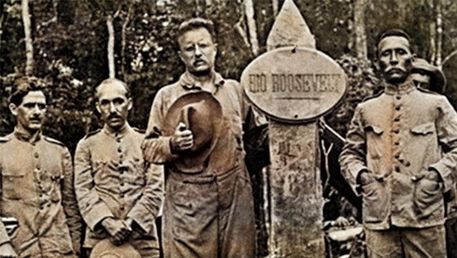 Theodore Roosevelt standing beside the marker reading RIO ROOSEVELT, with officers of the Rondon expedition, 1914